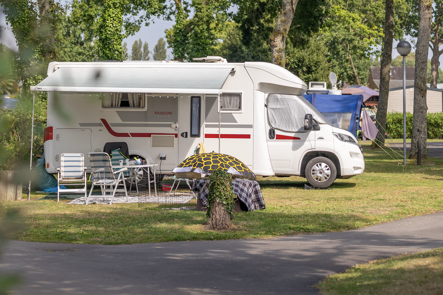 Wohnmobil auf einem Campingplatz mit Vorzelt, Stühlen und Tisch, daneben ein Sonnenschirm im Schatten der Bäume.