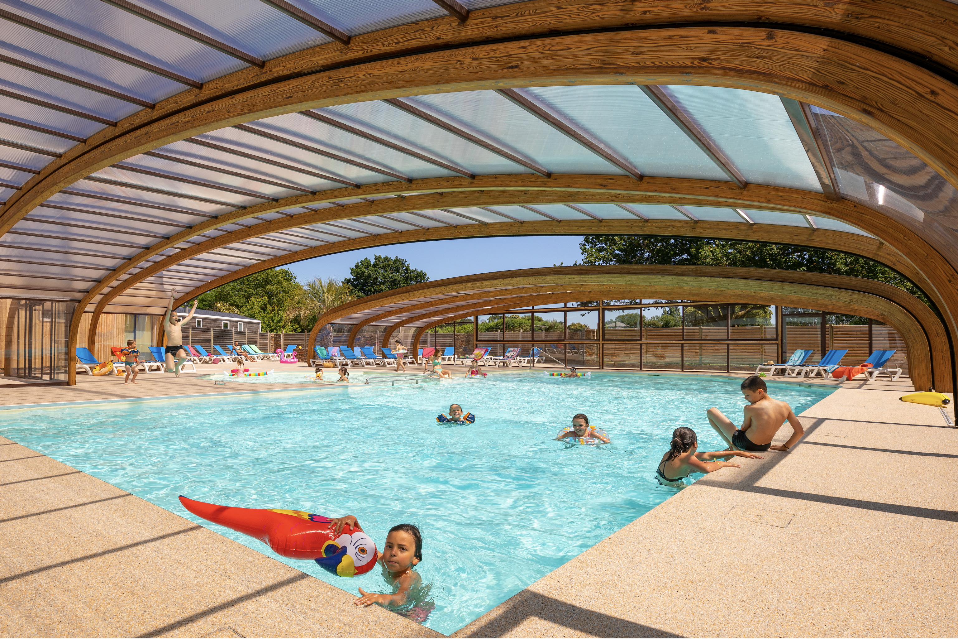 Innenpool mit türkisfarbenem Wasser, Liegen und Sonnenschirmen unter einer transparenten Überdachung, Kinder und Erwachsene im Becken.