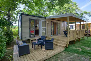 Mobilheim mit Holzterrasse, Sitzgruppe und Markise am Eingang, umgeben von Bäumen und Rasen im Campingbereich.