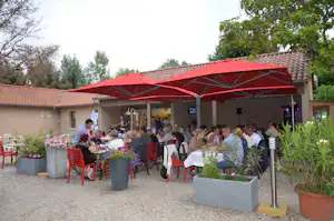 Terrasse am Campingplatz mit langen Tischen, roten Sonnenschirmen und Sitzplätzen vor einem Gebäude, umgeben von Pflanzen und Kiesweg.