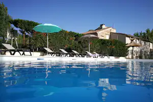 Blauer Pool mit Liegen, Sonnenschirmen und Zaun im Garten; dahinter ein Steinhaus mit Ziegeldach.