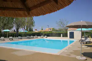 Pool im Campingbereich mit Sonnenschirmen, Liegestühlen und Blick auf Hecken, Bäume und Gebäude im Hintergrund.