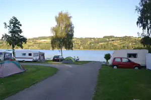 Campingplatz am See: Zelt und Wohnwagen entlang eines gepflasterten Wegs, rote und schwarze Autos, im Hintergrund grünes Ufer.