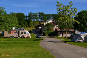 Wohnmobil und Zelt auf einem Campingplatz, gepflasterter Weg zur Rezeption, Bäume und Rasen im Hintergrund.