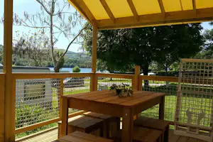 Holzterrasse mit Tisch und Bänken unter gelbem Sonnendach, Blick auf See, Bäume und grünes Ufer.