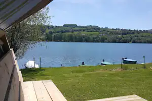 Blick vom Holzdeck auf den See: Zelt am Ufer, Rasenfläche, Bäume und grüne Hügel im Hintergrund.
