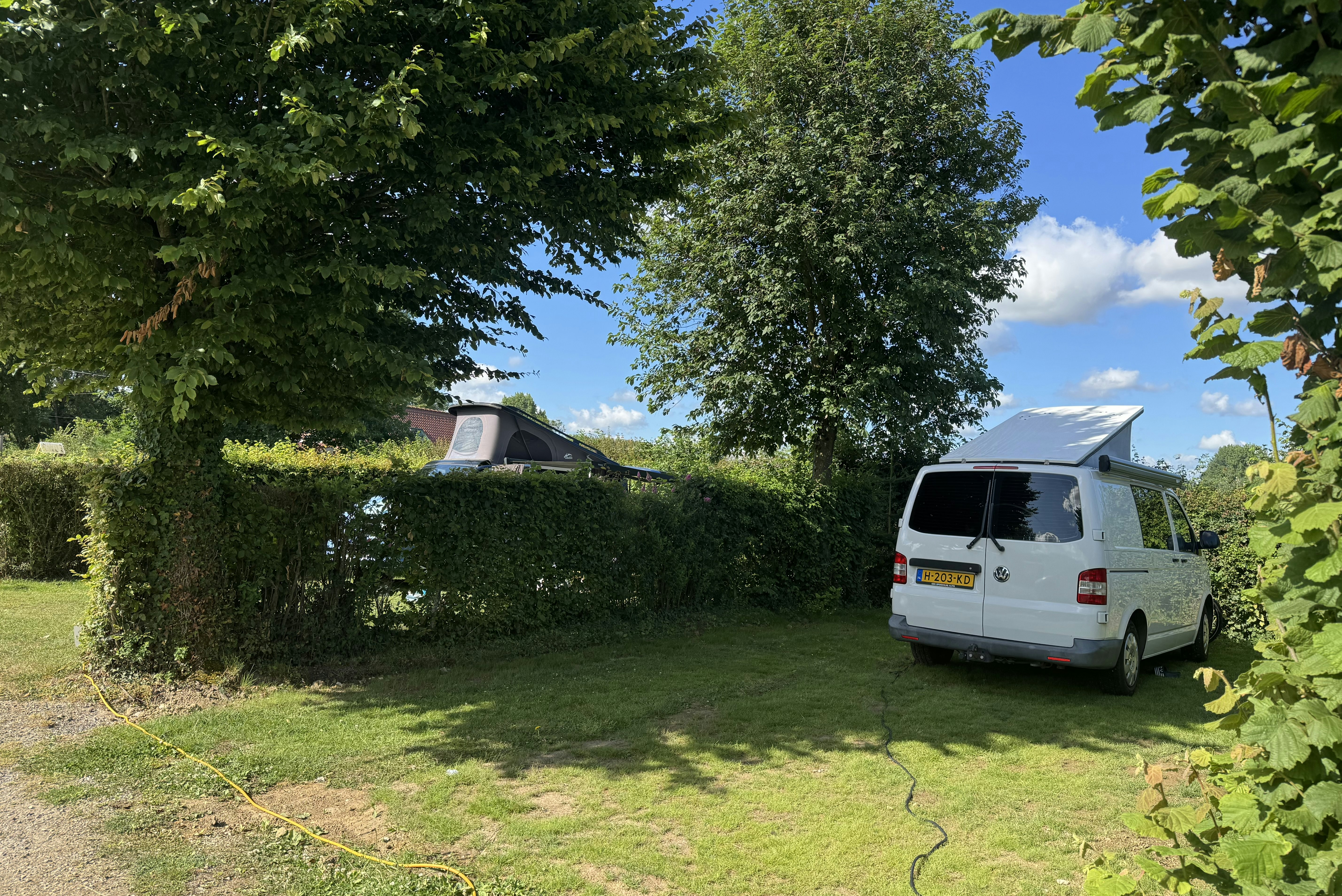 Weißer Camping-Transporter steht auf dem Stellplatz neben grünen Hecken und Bäumen, im Hintergrund ein Wohnwagen.