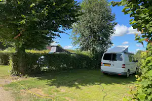 Weißer Camping-Transporter steht auf dem Stellplatz neben grünen Hecken und Bäumen, im Hintergrund ein Wohnwagen.