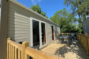 Bungalow mit Holzterrasse und Holzzaun, daneben ein Tisch und Stühle vor großen Fenstern, im Grünen.