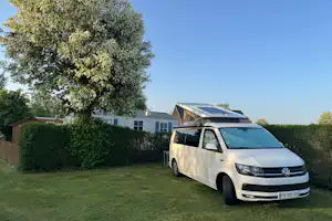 Weißes Wohnmobil auf einem Grasstellplatz am Campingplatz, mit Hecke, Baum und Gebäuden im Hintergrund.
