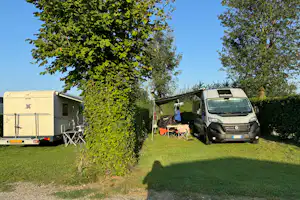 Wohnwagen und Campingbus auf einem Stellplatz, mit Tisch und Stühlen vor dem Fahrzeug, umgeben von Bäumen und Rasen.
