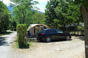 Auto und Zelt auf einem Campingstellplatz, neben einem Kiesweg und Büschen, unter großen Bäumen mit Blick auf Berge.