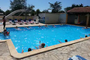 Blauer Camping-Pool mit mehreren Liegestühlen am Rand, dahinter ein Zaun und ein Gebäude mit rotem Ziegeldach.
