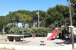 Spielbereich auf dem Campingplatz mit roter Rutsche, Federwippe, Sandfläche, Hecke und Bäumen im Hintergrund.