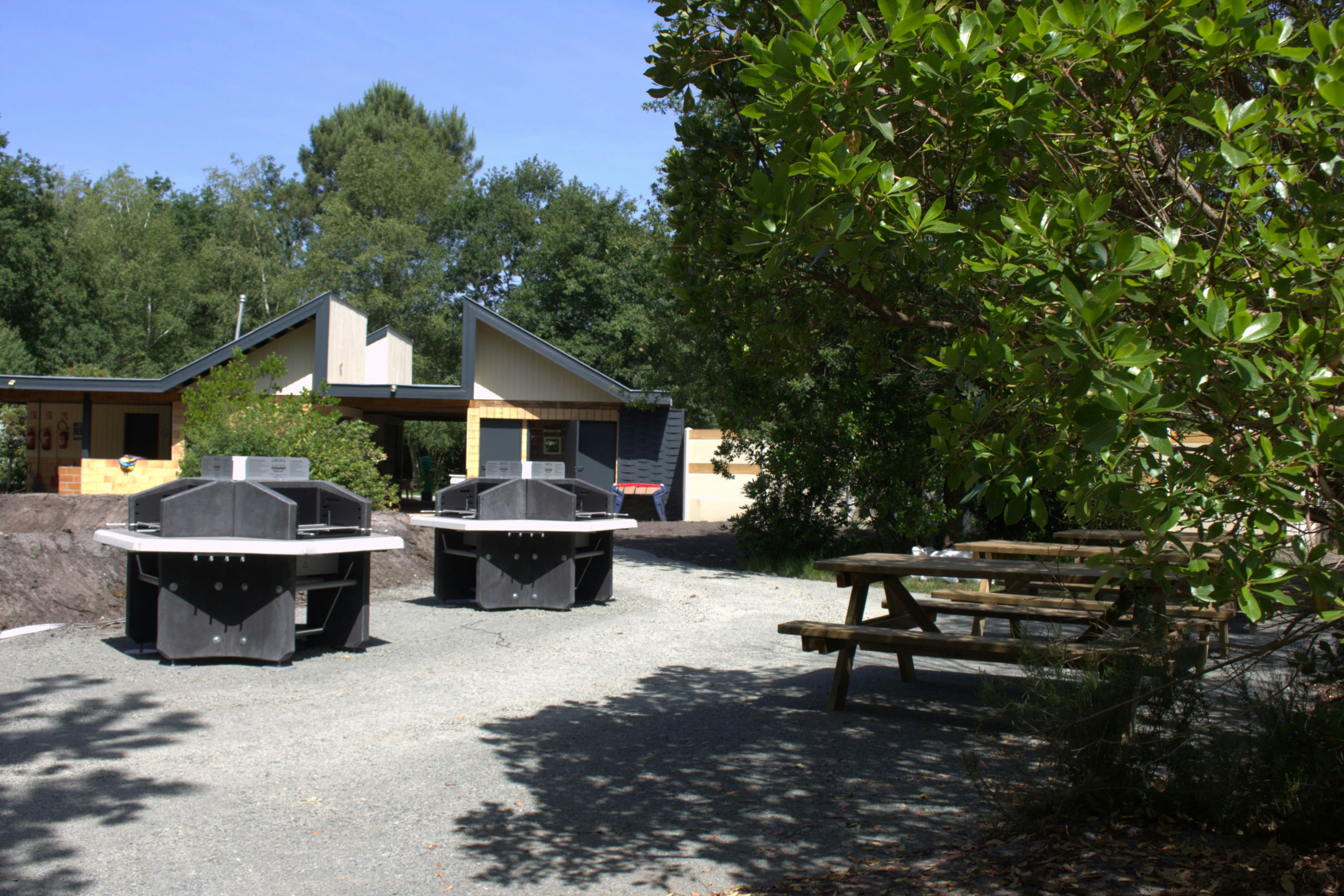Grillplatz mit zwei Grills auf gepflastertem Weg, daneben Sitzbank und Bungalows mit Terrasse im Grünen.
