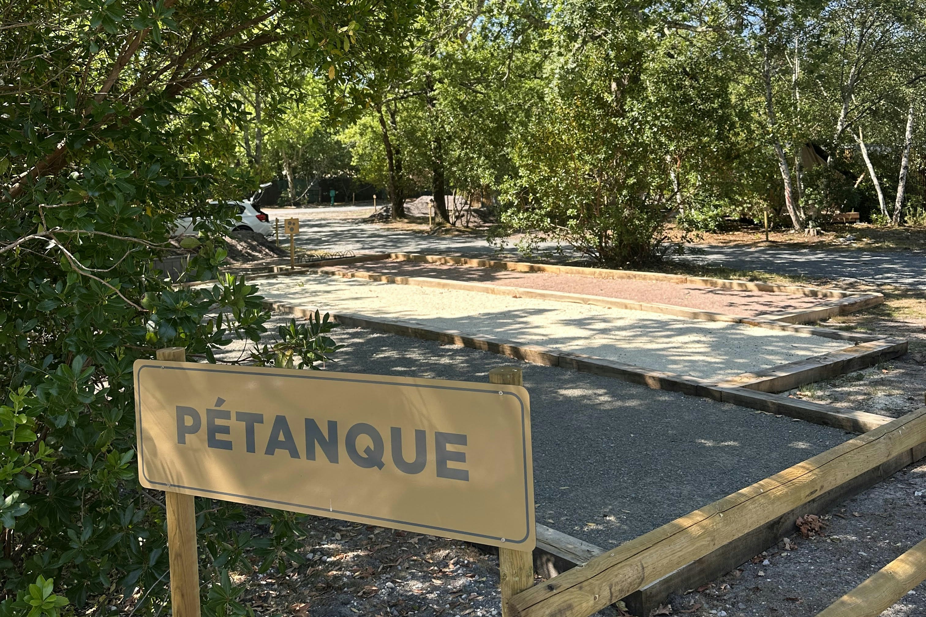 Schild „PÉTANQUE“ am Petanque-Spielplatz mit Kiesfläche, Holzgeländer und Bäumen im Hintergrund.