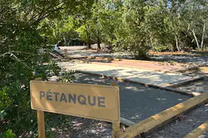 Schild „PÉTANQUE“ am Petanque-Spielplatz mit Kiesfläche, Holzgeländer und Bäumen im Hintergrund.