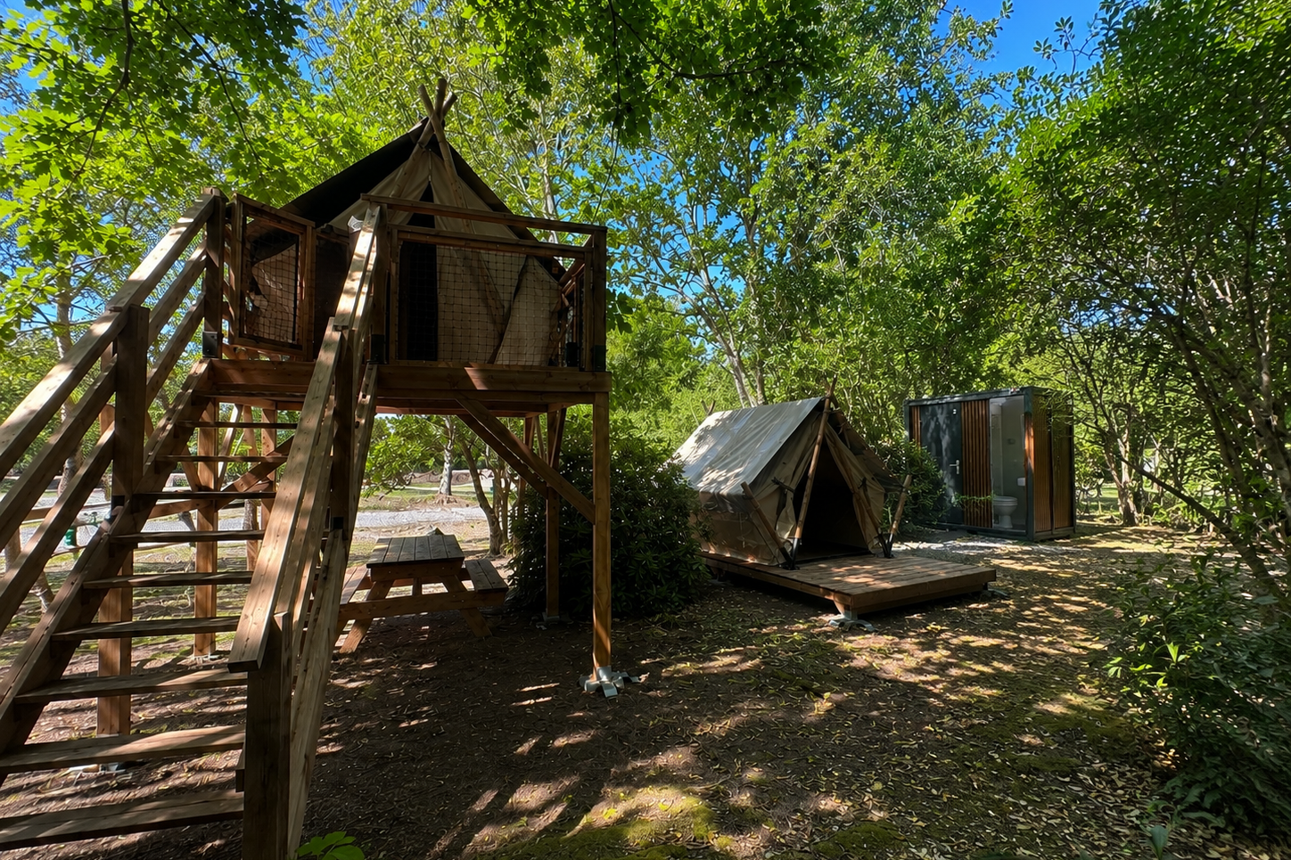 Holzplattform mit Treppe und Zelt auf einem Campingplatz im Grünen, daneben weitere Zelte und ein Weg zwischen Bäumen.