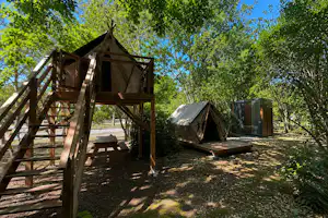 Holzplattform mit Treppe und Zelt auf einem Campingplatz im Grünen, daneben weitere Zelte und ein Weg zwischen Bäumen.