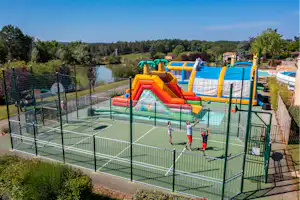 Kinder-Spielplatz mit Hüpfburg und Rutsche in einem eingezäunten Bereich, dahinter Bäume und ein kleiner See.