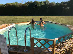 Campingpool mit zwei Kindern im Wasser, Holzgeländer am Beckenrand und grüner Wiese mit Bäumen im Hintergrund.