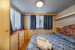 Bett im Wohnbereich eines Camping-Mobilehomes mit Vorhängen, Schrankwand und Deckenleuchte im Raum.