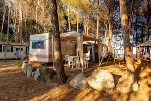Wohnwagen mit geöffneter Markise und Terrasse, daneben Campingstühle und Tisch im Wald zwischen Bäumen und Steinen.