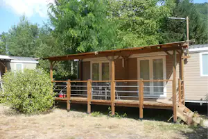 Holzbungalow mit überdachter Terrasse, Sitzgelegenheiten und großen Fenstern, umgeben von Büschen und Bäumen im Campingbereich.
