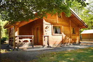 Holzhütte mit Veranda, Tisch und Bank, Fenster mit Vorhang; im Hintergrund Zelte und Bäume im Grünen.