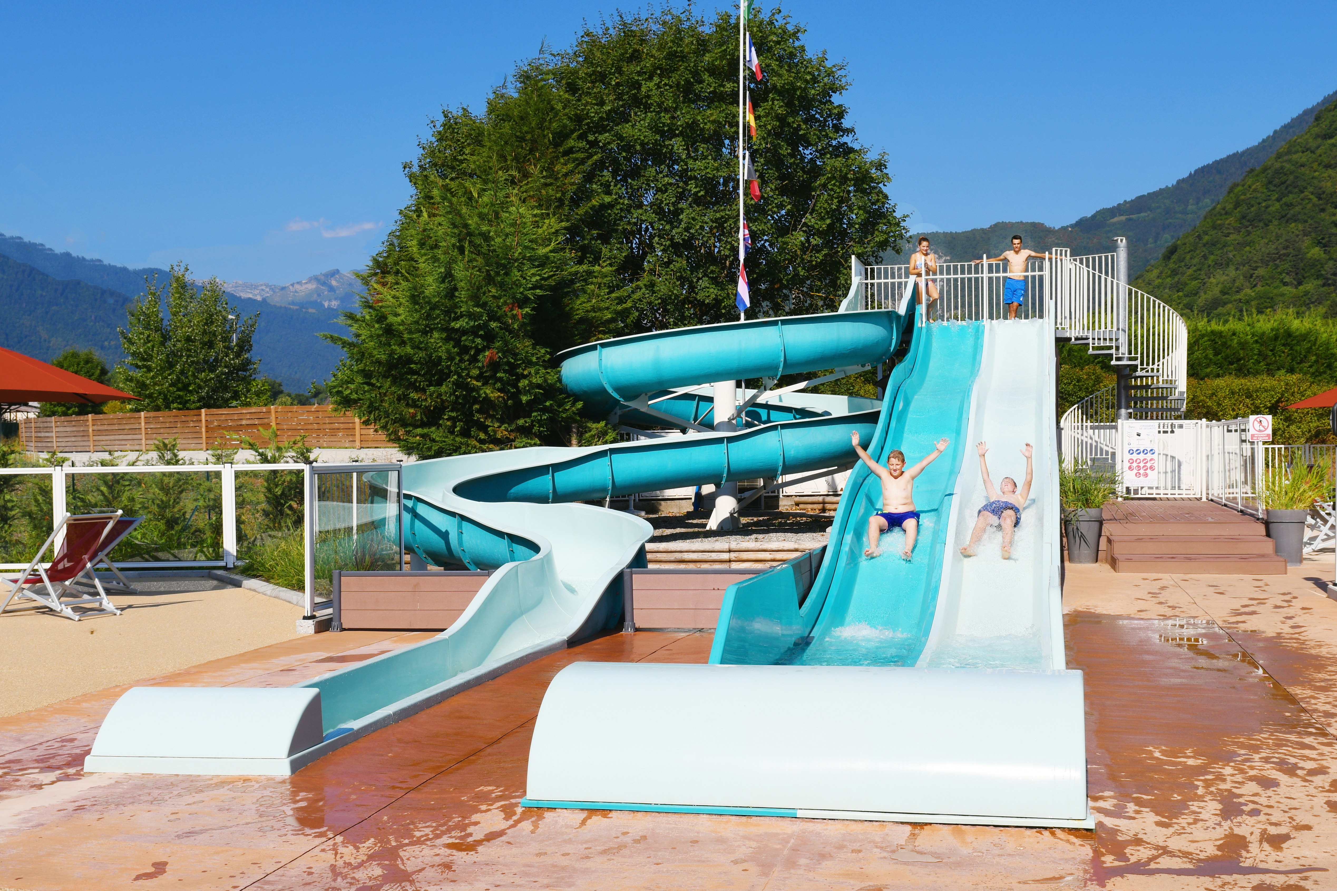 Wasserrutschen am Campingplatz mit türkisfarbenen Bahnen, Holzterrasse, Liegestühlen und Blick auf Berge und Bäume.