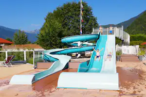 Wasserrutschen am Campingplatz mit türkisfarbenen Bahnen, Holzterrasse, Liegestühlen und Blick auf Berge und Bäume.