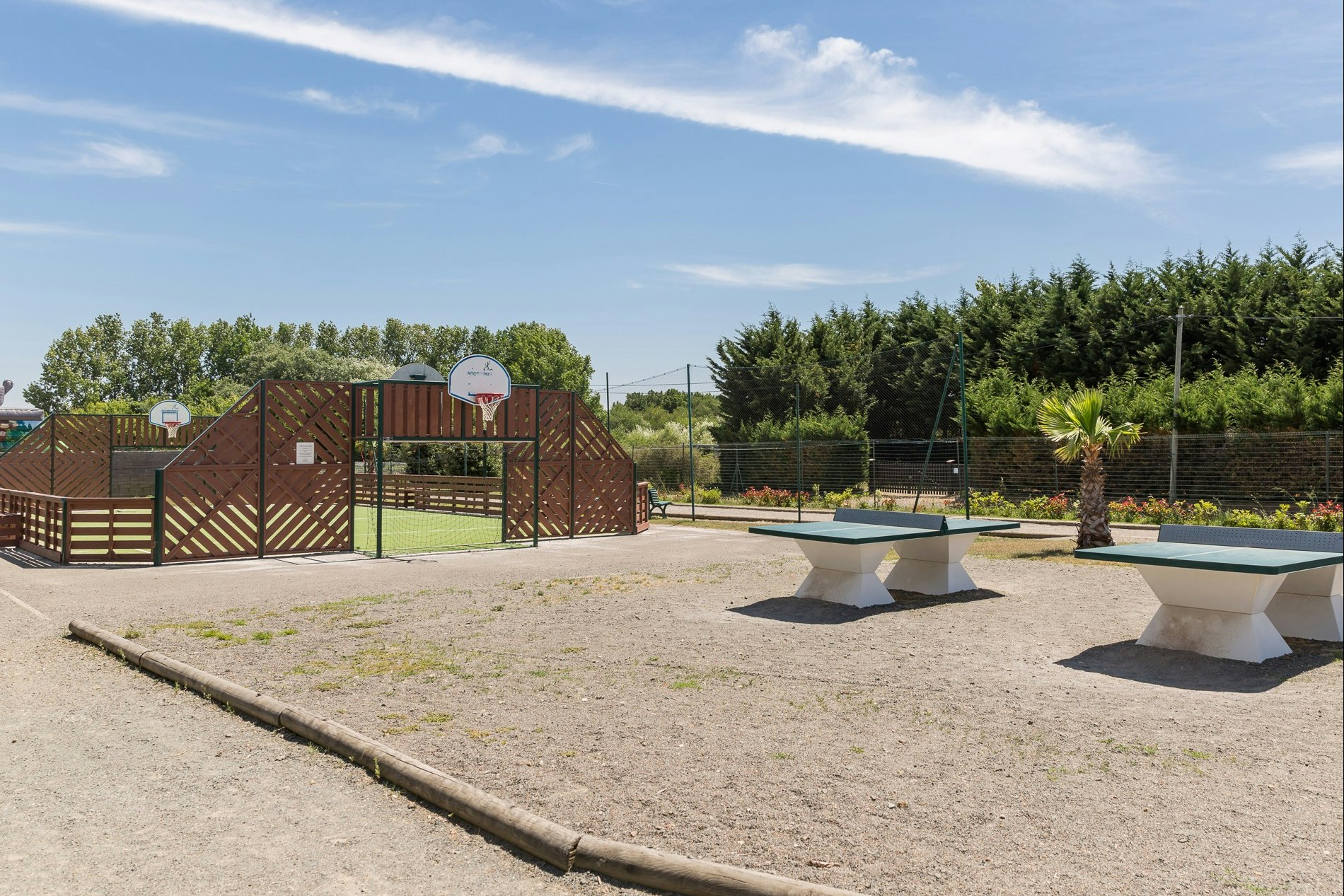 Campingplatz mit gepflasterter Fläche, Sitzbänken und Grillbereich vor einem Zaun, dahinter Bäume und ein Gebäude.