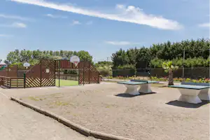 Campingplatz mit gepflasterter Fläche, Sitzbänken und Grillbereich vor einem Zaun, dahinter Bäume und ein Gebäude.