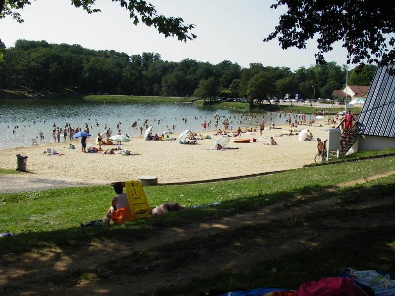 Badestrand am See mit Liegen und Sonnenschirmen, im Hintergrund Bäume und ein Campingbereich am Ufer.
