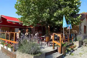 Terrasse mit Sitzgruppe und Pflanzen vor einem roten Gebäude, Bäume spenden Schatten auf dem Campingplatz.