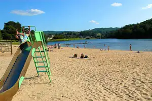 Badestrand am See mit Liegestühlen und Menschen im Hintergrund, im Vordergrund ein grüner Einstieg am Pool.
