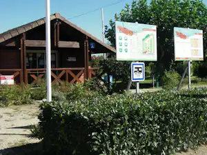 Holzhütten am Campingplatz mit Wegweisern und Schildern, Hecken und Kiesweg vor den Gebäuden.