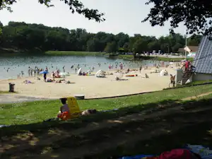 Badestrand am See mit vielen Liegen und Sonnenschirmen, im Hintergrund Bäume und ein Uferweg.