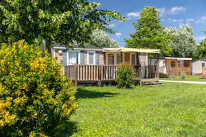 Mobilheim mit Terrasse, gelb-weißem Markisentuch und Holzgeländer im Grünen, umgeben von Bäumen und Rasen.