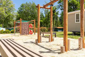 Spielplatz am Campingplatz mit Holzpfosten, Klettergerüst und Sandfläche, daneben ein Wohnwagen mit Terrasse und Zelt.