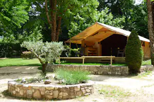 Campingzelt mit Holzveranda auf einem Stellplatz, umgeben von Bäumen, Hecken und einem kleinen Steinbeet.