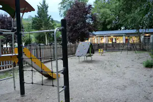 Spielplatz mit gelber Rutsche und Kletterelementen auf dem Campingplatz, dahinter Bäume und ein Gebäude mit beleuchteten Fenstern.