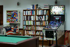 Bücherregal mit vielen Büchern, daneben Flipperautomat und Spielwand an der weißen Wand im Innenbereich.
