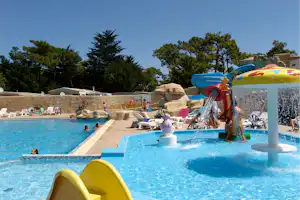 Familienpool mit Wasserspiel, Rutschen und Spritzfontäne, Liegen am Beckenrand und Bäume im Hintergrund.