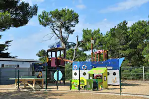 Bunter Spielplatz mit Zaun, Kletterturm und Rutsche, im Hintergrund hohe Kiefern und ein Gebäude am Campingplatz.