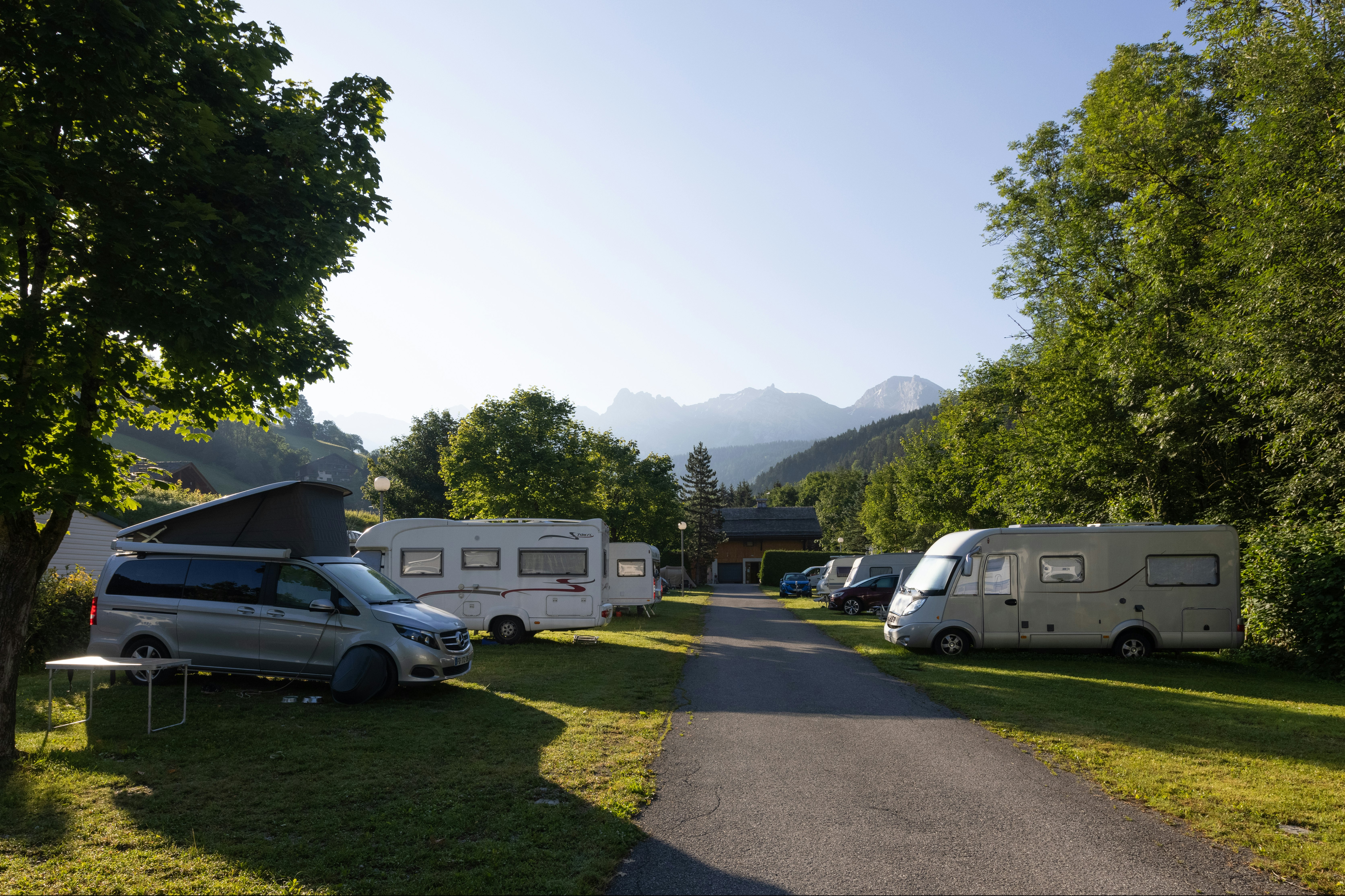 Campasun Camping Le Clos du Pin - Standplätze mit Wohnwagen und Wohnmobilen in der Abendsonne umgeben von Bäumen mit Blick auf die Berge