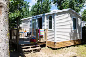 Weiße Mobilheim-Unterkunft mit Holzveranda, Tisch und Stühlen, bunten Decken und Blick auf Bäume im Campingbereich.