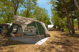 Grünes Campingzelt mit Vorzelt, Sitz- und Küchentisch auf Betonstellplatz, umgeben von Bäumen im Campingbereich.