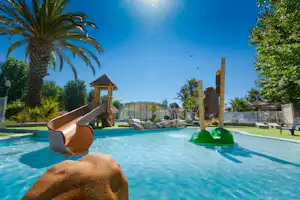 Pool am Campingplatz mit Palmen, Liegen und Spielbereich, im Hintergrund ein kleines Holzhaus und ein Glasdach.