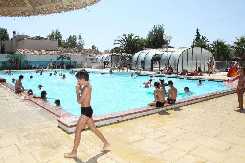 Außenpool auf einem Campingplatz mit Sonnenschirmen, Menschen im Wasser und einer überdachten Schwimmfläche im Hintergrund.